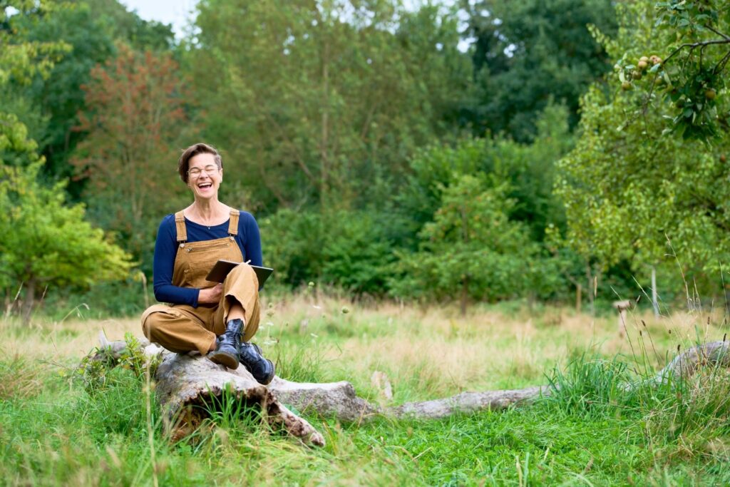 Sonja Stadje sitzt lachend auf einem Baumstamm inmitten von Obstbäumen