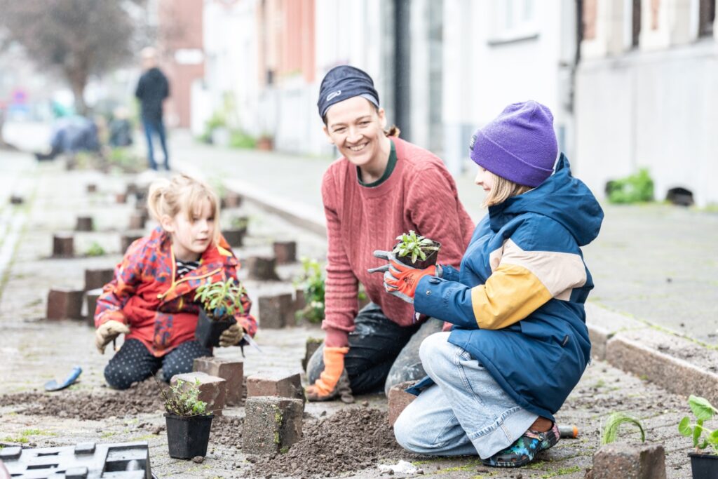 Eine Frau mit zwei Kindern macht beim Tegelwippen/Abpflastern mit und alle haben sichtlich Spaß beim Blumen pflanzen