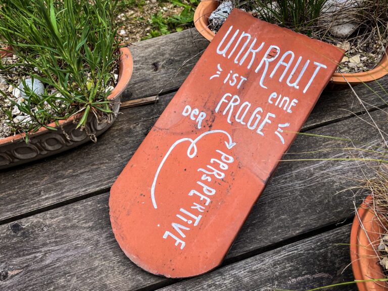 Ein Ziegel mit der AUfschrift "Unkraut ist eine Frage der Perspektive" liegt auf einem Holzdeck